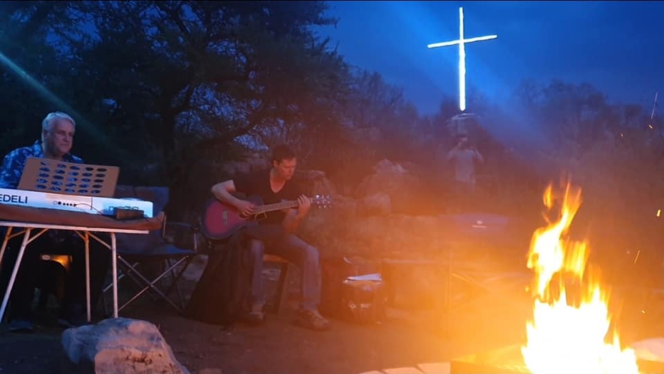 Worship around the fire at night with a glowing cross in the background.